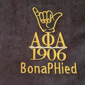 May include: A black towel with gold embroidery of a hand gesture, the letters "AΦA", the number "1906", and the words "Bona Fide".