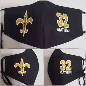 Op de afbeelding: Zwart gezichtsmasker met een gouden fleur-de-lis en het woord "Saints" geborduurd aan de ene kant en het nummer "32" met de naam "Mathieu" geborduurd aan de andere kant.