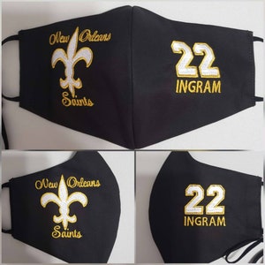 Op de afbeelding: Zwart gezichtsmasker met gouden borduurwerk. De linkerzijde heeft een fleur-de-lis en de tekst "New Orleans Saints". De rechterzijde heeft het nummer "22" en de tekst "INGRAM".