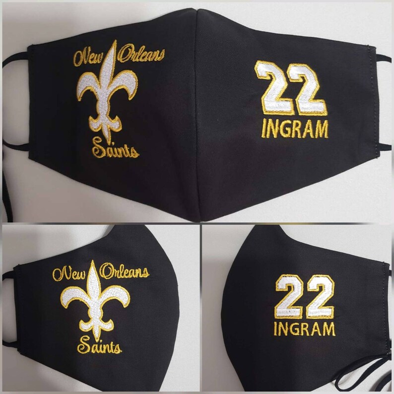 Op de afbeelding: Zwart gezichtsmasker met gouden borduurwerk. De linkerzijde heeft een fleur-de-lis en de tekst "New Orleans Saints". De rechterzijde heeft het nummer "22" en de tekst "INGRAM".