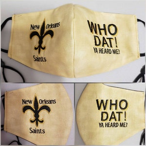 Op de afbeelding: Een beige gezichtsmasker met zwarte borduursels. Het masker heeft het logo van de New Orleans Saints en de tekst "WHO DAT! YA HEARD ME?"