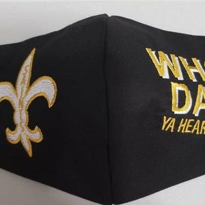 Op de afbeelding: Zwart gezichtsmasker met een gouden geborduurde fleur-de-lis aan de ene kant en de woorden "WHO DAT! YA HEARD ME?" aan de andere kant.