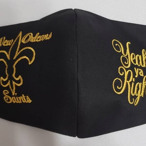 Op de afbeelding: Zwarte gezichtsmasker met gouden borduurwerk. Aan de ene kant staat het logo van de New Orleans Saints met de tekst "New Orleans Saints". Aan de andere kant staat de tekst "Yeah Ya Right!"