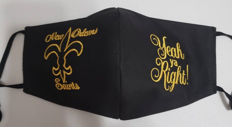 Op de afbeelding: Zwarte gezichtsmasker met gouden borduurwerk. Aan de ene kant staat het logo van de New Orleans Saints met de tekst "New Orleans Saints". Aan de andere kant staat de tekst "Yeah Ya Right!"