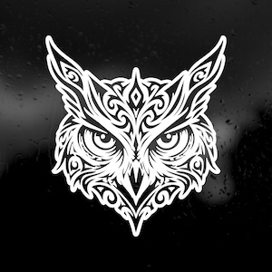 May include: A white vinyl decal featuring a stylised owl head with intricate, symmetrical tribal-inspired patterns. The design showcases sharp, pointed ear tufts, large focused eyes, and a pointed beak, all rendered in clean, bold white lines.