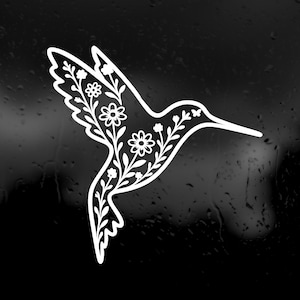 May include: A white vinyl decal of a hummingbird in flight, featuring a detailed internal pattern of flowers, leaves, and stems. The bird is depicted with its wings spread upwards and its long, thin beak pointing towards the right side of the frame.