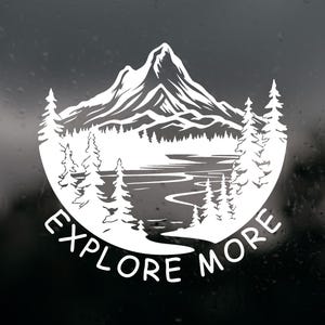 May include: A white vinyl decal featuring a detailed mountain range, a forest of pine trees, and a winding river. The design is circular in shape and includes the text EXPLORE MORE in a bold, sans-serif font curved along the bottom edge.