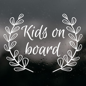 以下が含まれることがあります： 筆記体のフォントで「Kids on board」という文字が書かれた白いビニールステッカー。左右対称に描かれたスタイリッシュな葉の枝が中央の文字を囲むように上向きにカーブしており、月桂冠のようなデザインになっています。