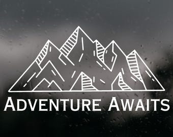 May include: A white vinyl decal featuring a line art illustration of a mountain range with hatched shading details. Below the mountain graphic, the text ADVENTURE AWAITS is printed in a clean, white, serif font. The design is applied to a dark surface.