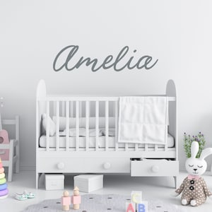 May include: A white cot with a white blanket and a grey name decal that says "Amelia" above it. The cot is in a nursery with a white rug, a stuffed bunny, and other toys.