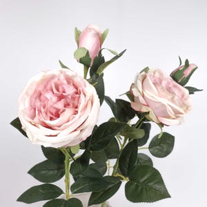 May include: A display of artificial roses, featuring open blooms and buds. The open flowers have soft pink petals with a touch of white, while the buds are a pale pink. Dark green leaves and stems complete the arrangement.