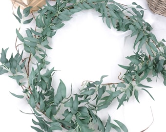 May include: A long garland of artificial greenery with small, light green leaves. The garland is arranged in a circle, creating a wreath-like shape.