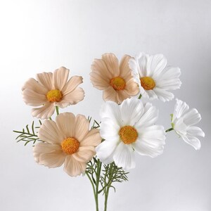 May include: A bunch of artificial cosmos flowers. The flowers have white and peach-coloured petals with yellow centres. Green stems and leaves are visible. The flowers are arranged against a white background.