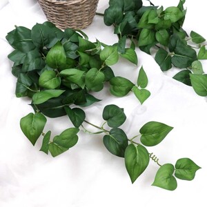 May include: A long strand of artificial green ivy with heart-shaped leaves. The ivy is draped over a white fabric surface.