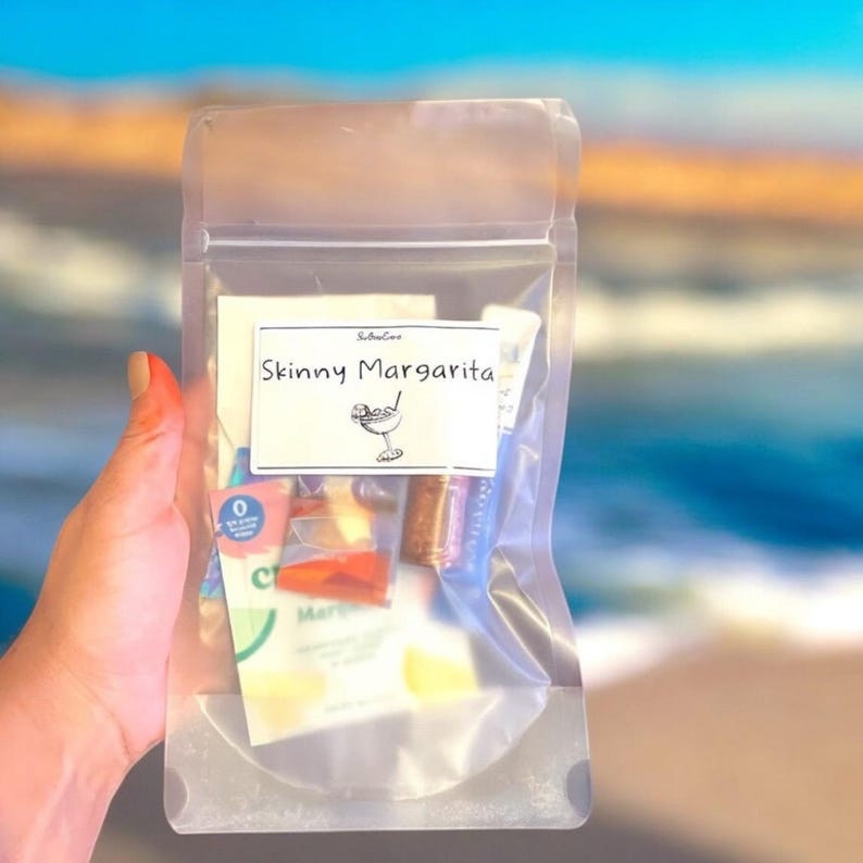 May include: A clear plastic bag with a label that says "Skinny Margarita" and contains several small packets of ingredients. The bag is held in a hand against a blurry background of a beach.