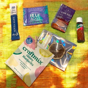 May include: A flat lay of various cocktail ingredients, including a packet of Skinny Margarita mix, a packet of blue agave, a packet of pink salt, a small bottle of chili flakes, and a dried lime slice.