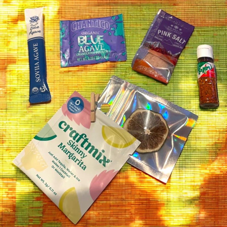 May include: A flat lay of various cocktail ingredients, including a packet of Skinny Margarita mix, a packet of blue agave, a packet of pink salt, a small bottle of chili flakes, and a dried lime slice.