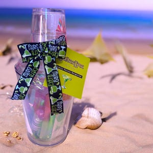 May include: A clear plastic cocktail shaker with a black and green ribbon tied around it. The shaker is filled with colorful cocktail ingredients. A green and yellow card with the text "SweetieLime Simply Skinny Margarita" is attached to the ribbon.