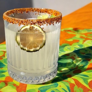 May include: A clear glass with a rim of chili powder and a lime wedge in a refreshing white drink. The glass is sitting on a colorful patterned surface.