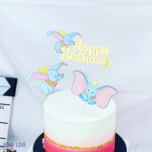Puede incluir: Un pastel blanco con glaseado rosa y dorado decorado con tres adornos de pastel de Dumbo el elefante y un adorno dorado de "Feliz cumpleaños".