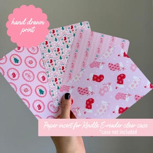 May include: A collection of pink paper inserts for a Kindle e-reader clear case, featuring various Christmas-themed patterns. Designs include candy canes, Christmas trees, stockings, and bows. The text "hand drawn print" is visible, along with "Paper insert for Kindle E-reader clear case *case not included."