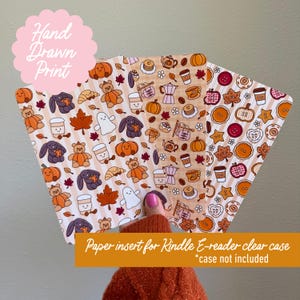 May include: Three rectangular paper inserts for a Kindle e-reader clear case featuring repeating autumn-themed patterns. The designs include illustrations of pumpkins, teddy bears, ghosts, coffee cups, maple leaves, and pastries in orange, brown, and white.
