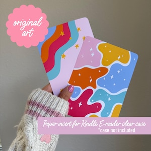 May include: Two colourful paper inserts for a Kindle e-reader clear case. One insert features a design with orange, pink, and blue wavy shapes with white stars. The other has a rainbow wave design with yellow stars. The text "original art" is in a pink circle.