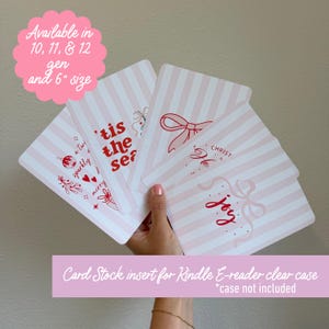 May include: Pink and white striped card stock inserts for a Kindle e-reader clear case. The cards feature holiday-themed text and illustrations in red, including bows, ornaments, and the words "Joy" and "'Tis the Season."