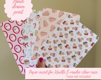 May include: A hand holding four decorative paper inserts for a Kindle e-reader clear case. The inserts feature festive patterns with illustrations of sweaters, ornaments, coffee cups, and candy canes. The text "Hand drawn print" and "Paper insert for Kindle E-reader clear case" are visible.