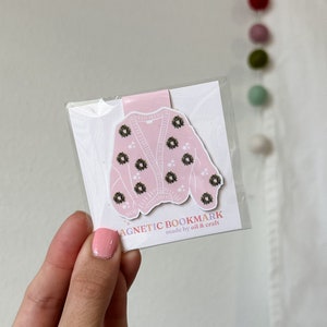 May include: Pink magnetic bookmark shaped like a cardigan with a wreath pattern. The bookmark says "Magnetic Bookmark made by oil & craft".