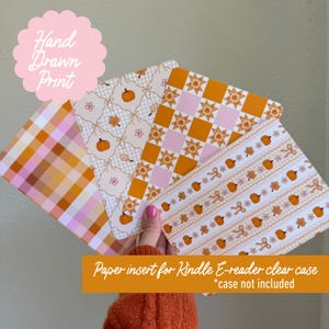 May include: Three rectangular paper inserts for a Kindle e-reader clear case. The designs feature orange pumpkins, autumn leaves, and flowers in patterns of plaid, checkered squares, and horizontal stripes with a cream and orange color palette.