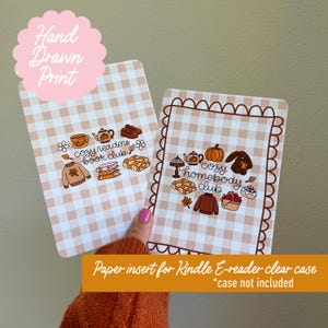 May include: Two rectangular paper inserts for a Kindle E-reader clear case featuring a beige and white gingham pattern. One insert displays the text cozy readers book club with illustrations of a sweater, books, and tea. The other shows cozy homebody club with a pumpkin, rabbit, and basket.