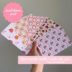 May include: A hand-drawn print of paper inserts for a Kindle e-reader clear case. The inserts feature various patterns with pumpkins, bats, and other fall-themed designs in orange, pink, and white. The text "hand drawn print" is visible.