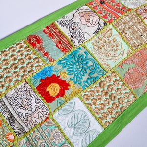 May include: A green patchwork table runner with embroidered floral designs in shades of green, blue, red, and gold. The runner is made of various fabrics and has a fringed edge.