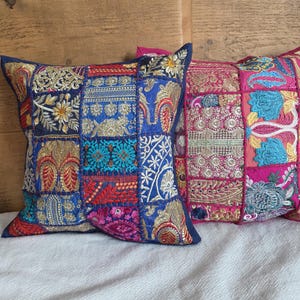 Puede incluir: Dos cojines decorativos con diseños de patchwork. Uno presenta un fondo azul con detalles bordados en rojo, dorado y turquesa. El otro cojín tiene un fondo rosa con bordados dorados y turquesas. Ambos cojines son cuadrados.
