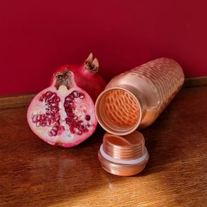 May include: A copper water bottle with a hammered finish and a matching lid. The bottle is open and next to a halved pomegranate.