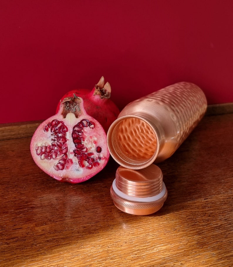 May include: A copper water bottle with a hammered finish and a matching lid. The bottle is open and next to a halved pomegranate.