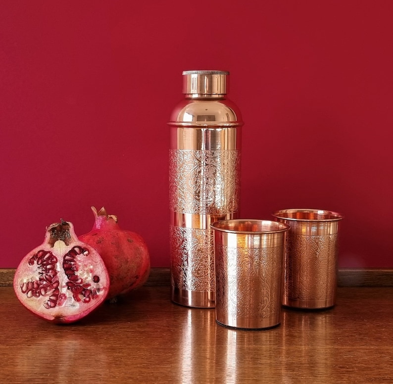 K&ouml;nnte beinhalten: Drei Kupfertrinkgef&auml;&szlig;e, zwei Tassen und eine hohe Flasche, mit einem verzierten gravierten Design. Die Flasche hat einen Schraubverschluss. Die Tassen und die Flasche stehen auf einer Holzoberfl&auml;che.