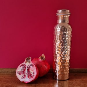May include: A copper water bottle with a hammered finish and a silver lid, next to two halves of a red pomegranate on a wooden surface.