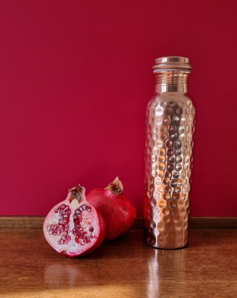 May include: A copper water bottle with a hammered finish and a silver lid, next to two halves of a red pomegranate on a wooden surface.