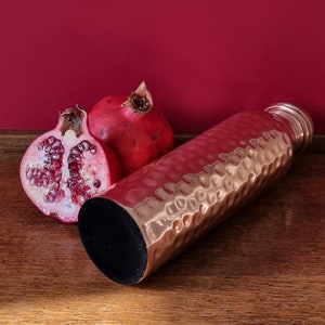May include: A hammered copper water bottle with a black lid, next to two halves of a red pomegranate on a wooden surface.
