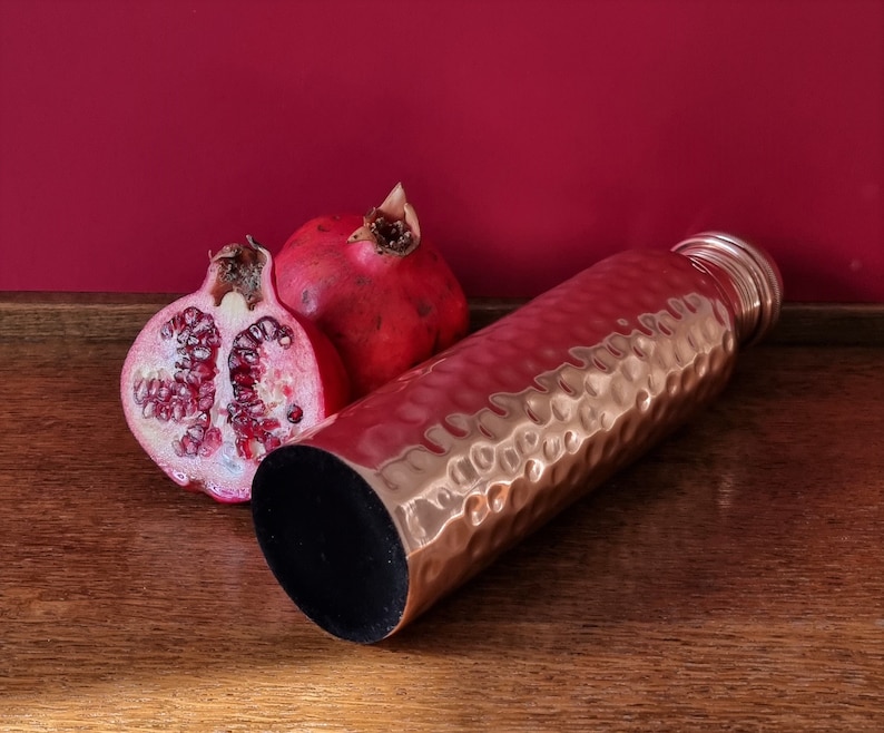 May include: A hammered copper water bottle with a black lid, next to two halves of a red pomegranate on a wooden surface.