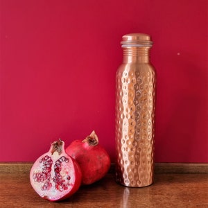 May include: A copper water bottle with a hammered finish and a silver lid, next to two halves of a red pomegranate on a wooden surface.