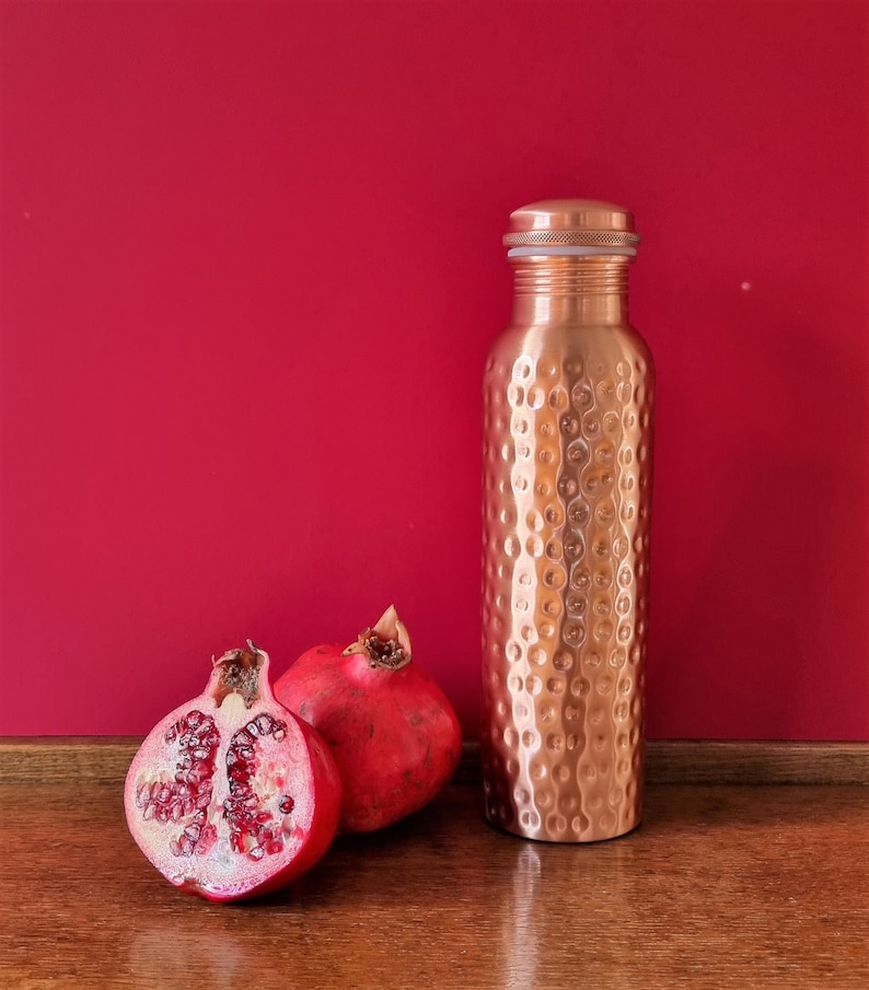 May include: A copper water bottle with a hammered finish and a silver lid, next to two halves of a red pomegranate on a wooden surface.