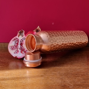 May include: A copper water bottle with a hammered finish and a matching lid. The bottle is lying on a wooden surface next to a half of a pomegranate.