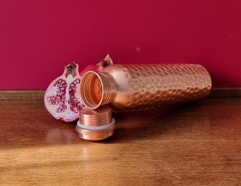 May include: A copper water bottle with a hammered finish and a matching lid. The bottle is lying on a wooden surface next to a half of a pomegranate.
