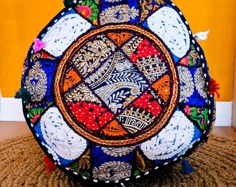 May include: Circular patchwork pouffe with a striking design. The centre showcases an intricate pattern in orange, red, and gold, framed by blue fabric and white sections. The edges are embellished with colourful tassels. This decorative item adds a bohemian flair to any room.