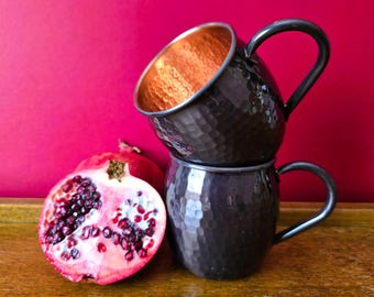 May include: Two black hammered metal mugs with copper interiors are stacked on top of each other. A halved pomegranate with visible seeds sits next to the mugs. The background is a solid magenta color, and the mugs are on a wooden surface.