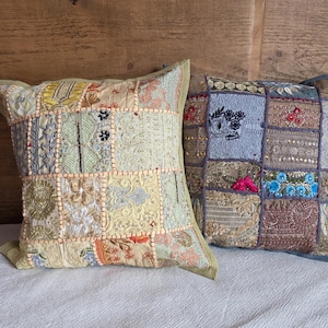 May include: Two decorative throw pillows with patchwork designs. The pillows feature a variety of fabrics in shades of beige, green, blue, and pink. Each pillow has a different pattern, with embroidered and textured details. The pillows are placed on a white surface.