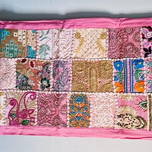 May include: A pink patchwork table runner with embroidered floral designs and beaded accents. The runner is made of various fabrics in shades of pink, beige, and brown.